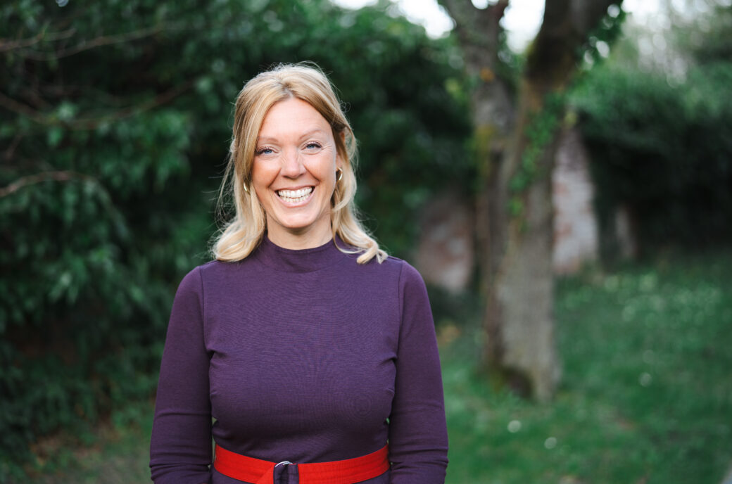Steffi, ein Mindful-Leadership-Coach der Framechangers™, lächelt in einem lila Kleid mit rotem Gürtel vor grünem Hintergrund.