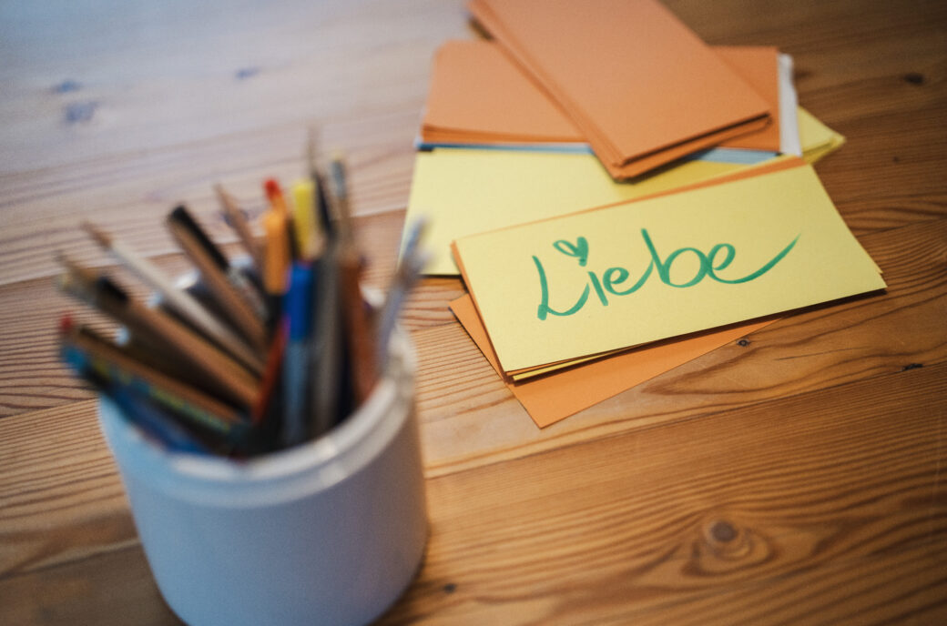 Buntstifte in Dose und Zettel mit "Liebe" Schriftzug auf Holztisch.