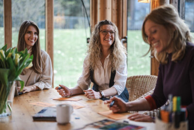 Team- & Organisationsentwicklung drei lächelnde Frauen sitzen am Tisch mit Stiften und Papier vor sich