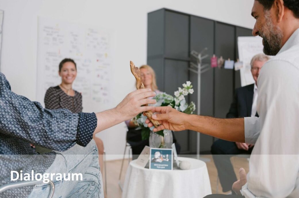 Zwei Hände reichen sich einen hölzernen Stab vor einer Gruppe von Personen; Text "Dialograum"